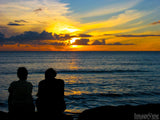 a couple sits by the ocean and watches the sunset in the background a couple sits by the ocean and watches the sunset in the background