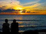 a couple sits by the ocean and watches the sunset in the background